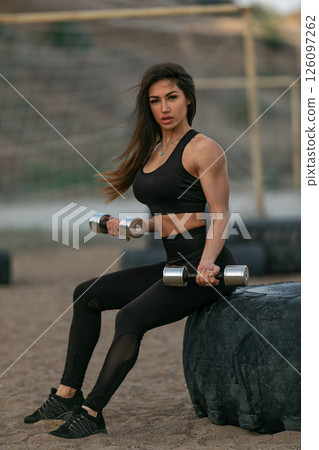 determined woman with long hair is performing strength training using dumbbells while seated on a large tire. She is dressed in black athletic wear at an outdoor gym setting. determined woman with long hair is performing strength training using dumbbells while seated on a large tire. She is dressed in black athletic wear at an outdoor gym setting. 126097262