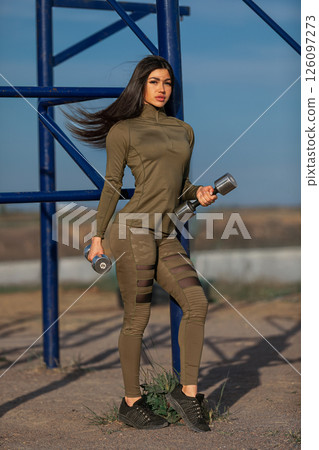 confident woman showcases her fitness style as she stands holding dumbbells at an outdoor gym surrounded by mountains. The clear blue sky adds to the vibrant atmosphere. 126097273