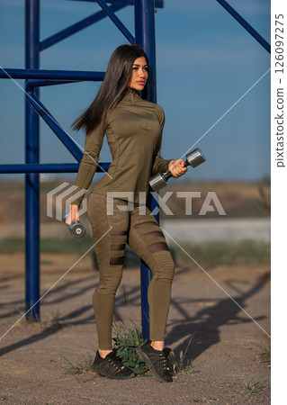 woman in olive green athletic attire holds dumbbells while standing confidently outdoors. The background features blue workout equipment and the day is bright and clear. woman in olive green athletic attire holds dumbbells while standing confidently outdoors. The background features blue workout equipment and the day is bright and clear. 126097275