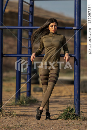 young woman stands elegantly at a playground wearing fitted athletic clothing. The sun shines brightly as she exudes confidence. The background features fitness equipment. young woman stands elegantly at a playground wearing fitted athletic clothing. The sun shines brightly as she exudes confidence. The background features fitness equipment. 126097284