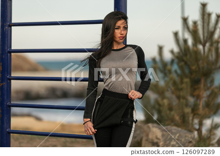 confident woman stands near outdoor gym equipment at the beach dressed in athletic wear. The day is sunny and calm highlighting her fitness routine amid nature's beauty. 126097289