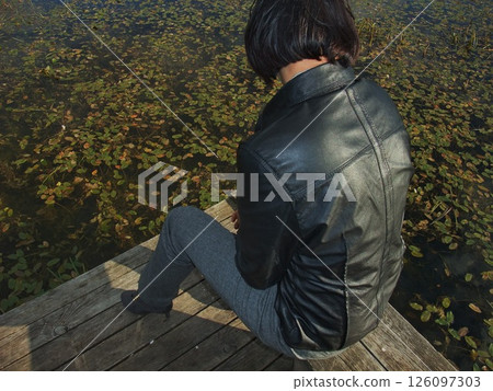 A person sits quietly on a rustic dock, facing a tranquil pond. The water is partially covered with green lily pads, creating a serene atmosphere on a sunny day. 126097303