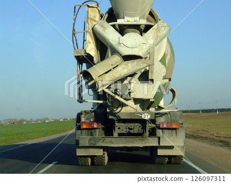 A large concrete mixer truck drives down a rural road, showcasing its heavy machinery and equipment used for construction, with fields visible in the background. 126097311