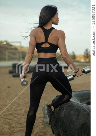 Woman in black athletic wear lifts dumbbells while standing on a tire at an outdoor gym. The sun shines brightly in the background highlighting her fitness and dedication. 126097321