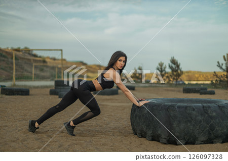 woman in workout attire is pushing a large tire on a sandy outdoor fitness area. The scene conveys strength and determination in a natural setting under a clear sky. 126097328