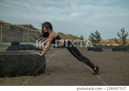 woman in athletic wear executes push ups on a large tire at an outdoor workout area. The setting features a sandy ground and distant hills under a cloudy sky ideal for fitness activities. 126097337
