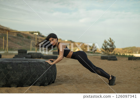 fit young woman engages in push up exercises using large tire as support in sandy outdoor fitness area surrounded by hills under a cloudy sky.. fit young woman engages in push up exercises using large tire as support in sandy outdoor fitness area surrounded by hills under a cloudy sky.. 126097341