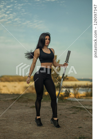 woman stands in athletic attire holding a barbell while showcasing her strength and fitness. The natural setting features greenery and a distant body of water under a clear sky. woman stands in athletic attire holding a barbell while showcasing her strength and fitness. The natural setting features greenery and a distant body of water under a clear sky. 126097347