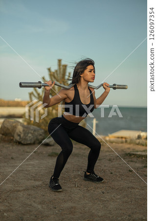 woman in athletic attire performs barbell squats in a natural setting with water in the background. She focuses intently on her form while exercising in a peaceful environment. 126097354