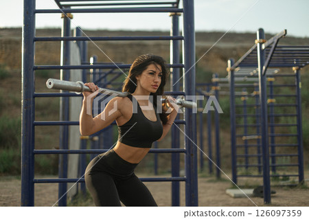 woman is engaged in a vigorous squat exercise using a barbell in an outdoor fitness space. The area is equipped with various workout structures surrounded by natural scenery. woman is engaged in a vigorous squat exercise using a barbell in an outdoor fitness space. The area is equipped with various workout structures surrounded by natural scenery. 126097359