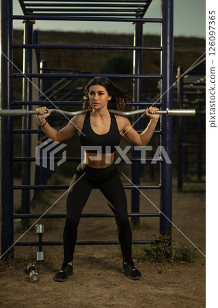fit woman performs a squat with a barbell in an outdoor gym setting as the sun sets in the background. She displays focus and determination in her workout. 126097365
