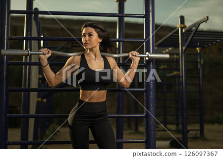 woman in athletic wear confidently lifts a barbell during a strength training session at an outdoor gym. The sun sets in the background creating a warm ambiance. 126097367