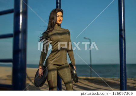 fit woman stands confidently at an outdoor gym near the beach holding weights while preparing for her workout. The calm ocean and bright sky create a perfect training environment. fit woman stands confidently at an outdoor gym near the beach holding weights while preparing for her workout. The calm ocean and bright sky create a perfect training environment. 126097369