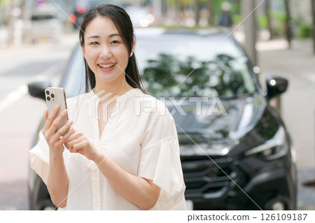 A young woman operating a smartphone in front of a car 126109187