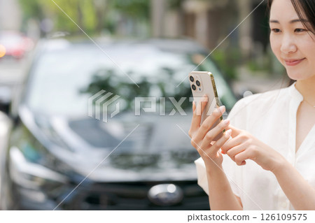 A young woman operating a smartphone in front of a car A young woman operating a smartphone in front of a car 126109575