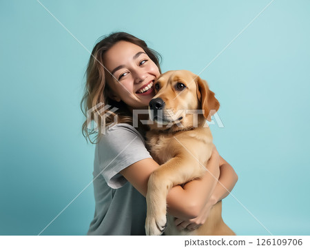 Woman embraces yellow Labrador retriever against a light blue background while smiling joyfully 126109706