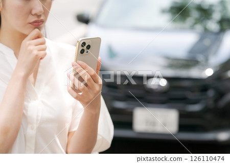 A young woman operating a smartphone in front of a car A young woman operating a smartphone in front of a car 126110474