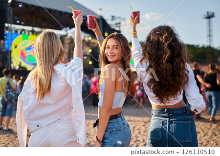 Group of girls at summer music festival on sandy beach celebrate, holding drinks, dancing. Happy women in casual outfits enjoy live concert, sunny seascape setting. Youth culture, friendship moments. 126110510