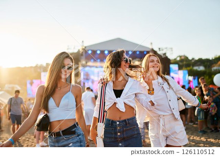 Three joyful young women stroll on sandy shore at sunset, friends laugh, dance at summer music beach fest. Trendy festival-goers in casual fashion enjoy live entertainment, golden hour vibes by sea. 126110512
