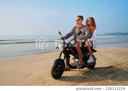 Couple enjoys beach ride on motorbike. Man drives motorcycle with woman on back. Ocean, sand, summer, sunglasses. Adventure travel, romantic journey, tropical holiday by sea. 126111224