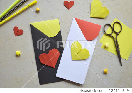 Handmade Crafting Process of a DIY Greeting Card with Origami Paper Hearts, Glue Application, Creative Stationery, Scissors, and Colorful Decorative Elements on a Light Artistic Background 126111249