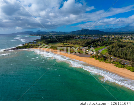 Aerial perspective of Kauai's coastline showing a sandy beach, turquoise waters, a parallel road, lush greenery, scattered buildings, and mountain peaks. Aerial perspective of Kauai's coastline showing a sandy beach, turquoise waters, a parallel road, lush greenery, scattered buildings, and mountain peaks. 126111674