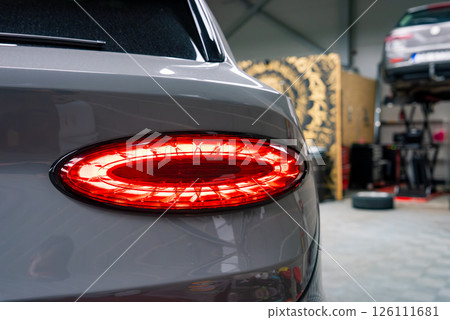 Rear left taillight of a Bentley Bentayga with oval red light design, parked indoors in a garage with a car lift, tools, and decorative panel visible. 126111681