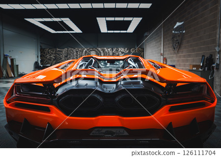 The image captures the rear end of a Lamborghini Revuelto indoors, showcasing its vibrant orange color, angular taillights, and distinctive exhaust system. 126111704
