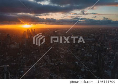 The image shows a sunset over London, highlighting the Shard and London Eye. The River Thames reflects the golden hues, contrasting with the dark cityscape. The image shows a sunset over London, highlighting the Shard and London Eye. The River Thames reflects the golden hues, contrasting with the dark cityscape. 126111709