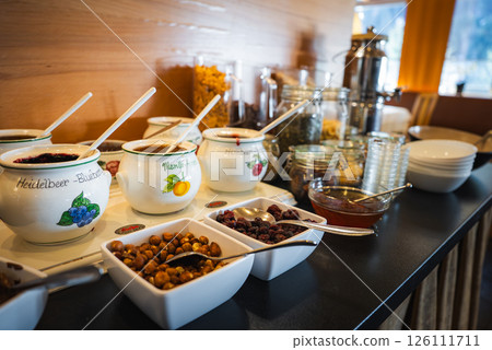 Breakfast buffet features labeled fruit jars, nuts, dried fruits, and cereals. Wooden decor and window reveal an alpine mountain landscape. 126111711
