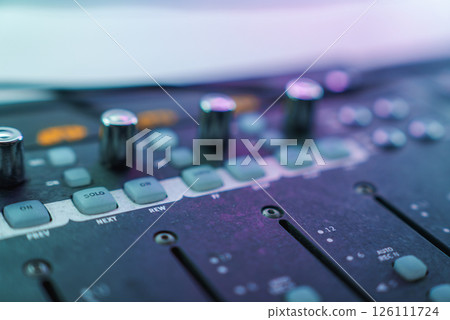 A detailed view of an audio mixing console with labeled knobs and buttons, illuminated by soft, colorful light, suggesting a music studio setting. 126111724