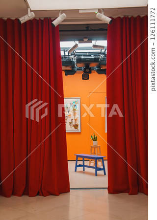 A modern room in London features vibrant red curtains, orange walls, a blue bench, and a wooden stool with a potted plant, under ceiling lights. A modern room in London features vibrant red curtains, orange walls, a blue bench, and a wooden stool with a potted plant, under ceiling lights. 126111772