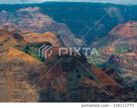 Waimea Canyon on Kauai Island, Hawaii, features rugged cliffs, deep valleys, and vibrant red, orange, and green hues with scattered vegetation. 126111773