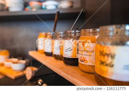 Wooden shelf with glass jars of honey and jam, labeled in Italian and German, with spoons inside. Blurred shelves in the background add depth. 126111775