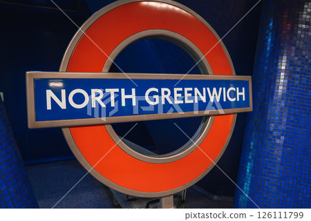 The iconic red and blue roundel sign for North Greenwich station is displayed against vibrant blue tiled walls in a modern London Underground setting. The iconic red and blue roundel sign for North Greenwich station is displayed against vibrant blue tiled walls in a modern London Underground setting. 126111799
