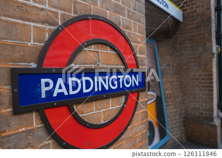 The iconic red and blue roundel sign of the London Underground at Paddington Station, mounted on a brick wall with background signage and poster details. 126111846