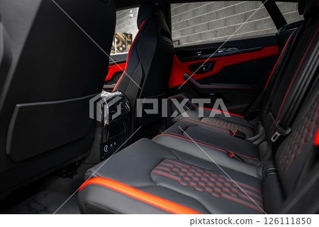 The interior of a Lamborghini Urus features black leather seats with red accents, intricate stitching, advanced controls, and a concrete wall in the background. 126111850