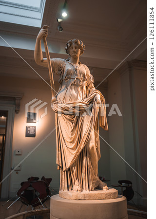 A marble statue of a draped female figure holding a staff, displayed indoors under soft lighting in a London museum with high ceilings and signage. A marble statue of a draped female figure holding a staff, displayed indoors under soft lighting in a London museum with high ceilings and signage. 126111854