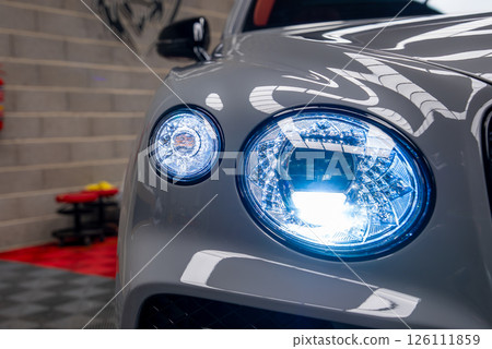 Front headlight assembly of a Bentley Bentayga with illuminated blue LED lights, gray exterior, tiled flooring, and a brick wall in a showroom. Front headlight assembly of a Bentley Bentayga with illuminated blue LED lights, gray exterior, tiled flooring, and a brick wall in a showroom. 126111859