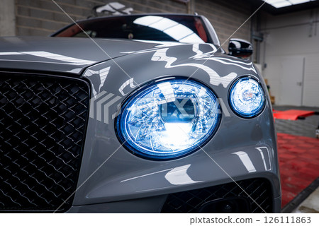 Close up view of a gray Bentley Bentayga showcasing circular LED headlights, black mesh grille, and polished surface in an indoor showroom setting. 126111863