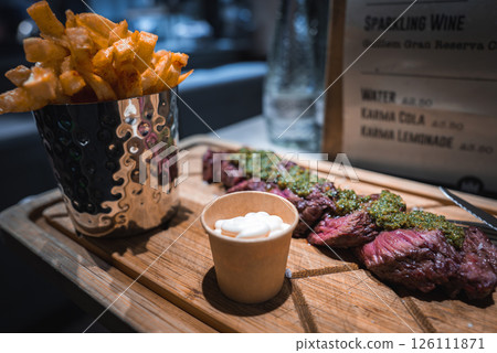 A wooden board holds sliced steak with green chimichurri and creamy sauce, paired with crispy fries in a metal cup. A menu board is visible in the background. 126111871