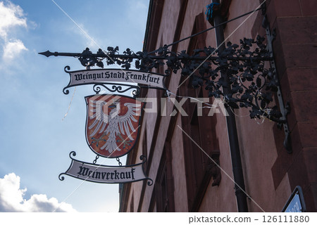 Decorative wrought iron sign reading Weingut Stadt Frankfurt and Weinverkauf, featuring a red shield with a white eagle, set against a blue sky. 126111880