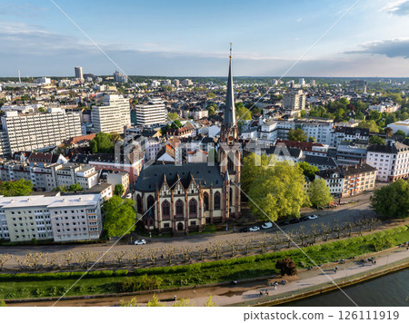 Aerial view of Frankfurt, Germany, highlighting Dreikonigskirche's Gothic spire, the Main River, tree lined pathways, and an expansive urban skyline. 126111919