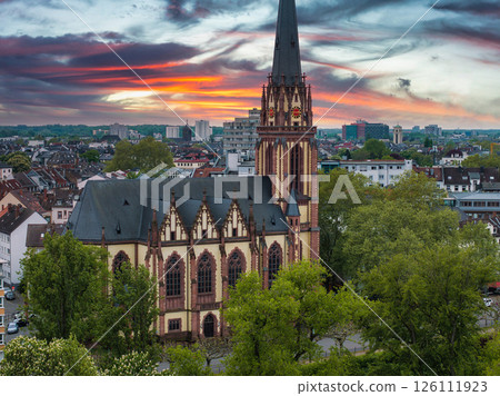 Aerial view of Dreikonigskirche in Frankfurt, Germany, with its Gothic spire, lush trees, urban surroundings, and a vibrant orange and purple sunset. Aerial view of Dreikonigskirche in Frankfurt, Germany, with its Gothic spire, lush trees, urban surroundings, and a vibrant orange and purple sunset. 126111923