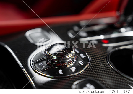 Close up view of a luxury car's center console with carbon fiber finish, metallic rotary knob, functional buttons, and red leather interior accents. 126111927