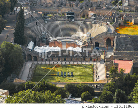 Aerial view of the Roman amphitheater in Pompeii, Italy, surrounded by ruins, stone walls, arches, greenery, and modern art installations in the foreground. Aerial view of the Roman amphitheater in Pompeii, Italy, surrounded by ruins, stone walls, arches, greenery, and modern art installations in the foreground. 126111963
