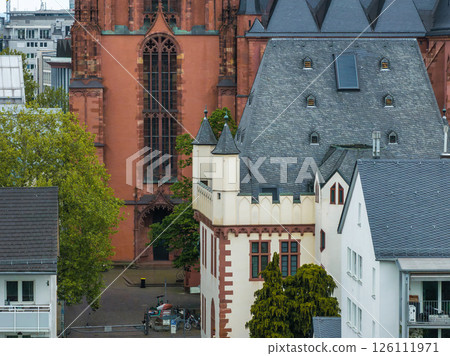 View of Frankfurt Cathedral with red sandstone facade, surrounded by traditional European buildings, greenery, and bicycles in the foreground. View of Frankfurt Cathedral with red sandstone facade, surrounded by traditional European buildings, greenery, and bicycles in the foreground. 126111971