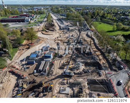 Expansive construction site in Riga with cranes, excavators, and temporary facilities, surrounded by urban and green spaces under a clear sky. 126111974