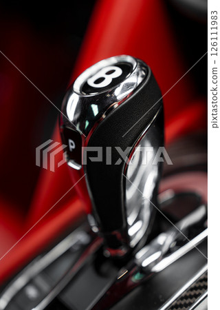 Close up view of a Bentley gear shifter in chrome and black leather, featuring the iconic B logo. Red interior accents are visible in the background. 126111983