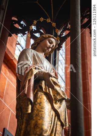 A detailed statue of a serene figure in golden robes with a halo of stars, set against red brick walls and stained glass in a Frankfurt church. 126111994